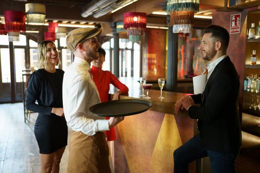 Male bartender with tray serving refreshing alcohol drinks on wooden counter for group of elegant people celebrating reunion in luxury bar