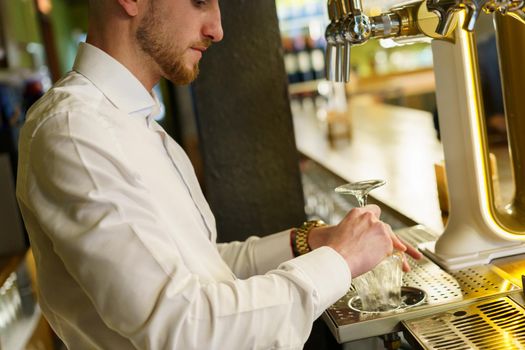 Unrecognizable male bartender pouring cold beer from tap into glass goblet during work in bar