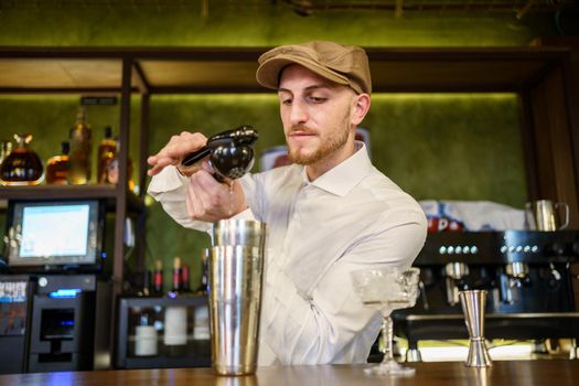 Bearded man in white shirt and cap squeezing lemon juice over shaker with cocktail while working behind counter in pub
