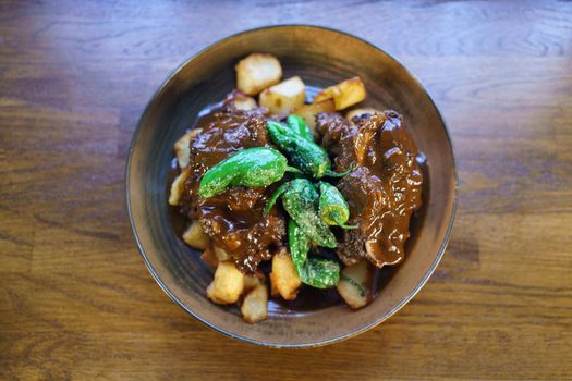 Top view of bull's tail, cooked and ready to eat in a Spanish restaurant, with tasty fried potato served with sauce and chili pepper in bowl on lumber restaurant table