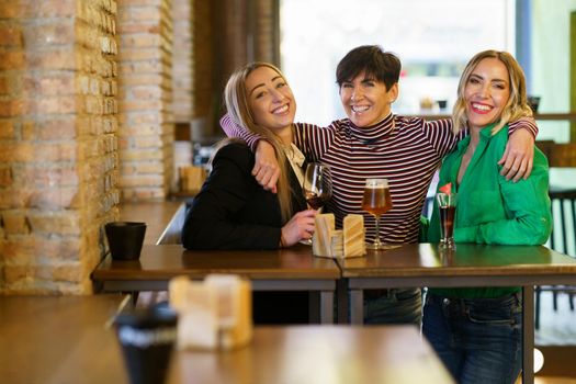 Cheerful group of female friends looking at camera with smile and embracing girlfriends behind table with alcohol during meeting in pub