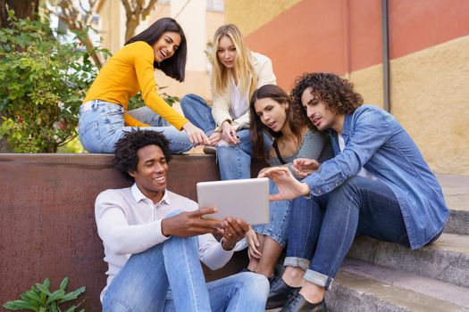 Group of multiethnic best friends gathering in city and watching funny video on tablet while spending time together