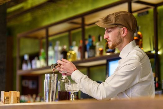 Bearded barman in white shirt and cap pouring liquor into shaker while preparing cocktail on counter in pub