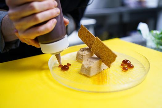Unrecognisable chef squeezing drops of jam onto a plate of foie micuit with crunchy croutons while working in a restaurant