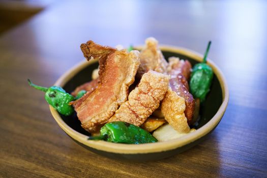 Torreznos, pieces of deep fried, meat with green hot peppers served in bowl on lumber table in restaurant