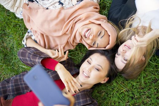 Top view of positive diverse female friends taking self portrait while lying together on green lawn on summer day in park