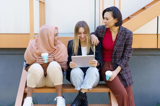 Trendy young multiracial female students in casual clothes drinking takeaway coffee and using tablet while sitting on stairs in city and preparing for exams