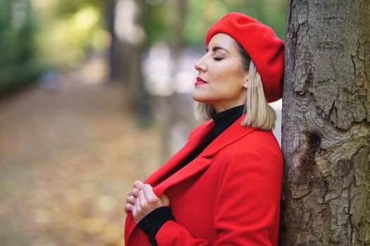 Side view of attractive female in stylish red coat with classic beret leaning on tree trunk with closed eyes in park on blurred background