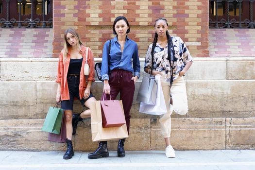 Full body of confident multiracial female friends with colorful shopping bags looking at camera while standing near building in city