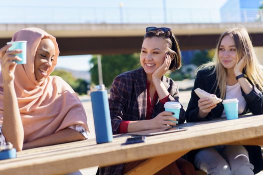 Positive young multiethnic female friends in casual clothes smiling while resting in outdoor cafe and drinking coffee from disposable cups