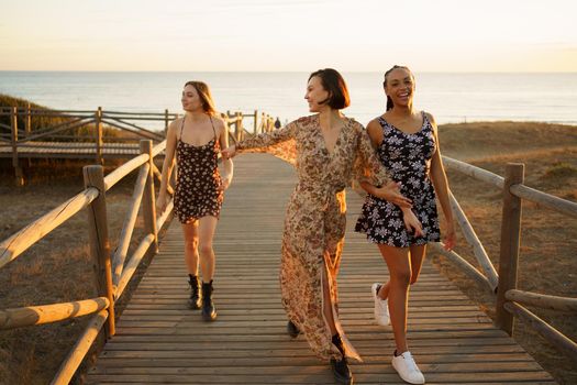 Full body of positive multiracial female friends strolling on wooden footbridge while spending time on seashore near sea on sunny day