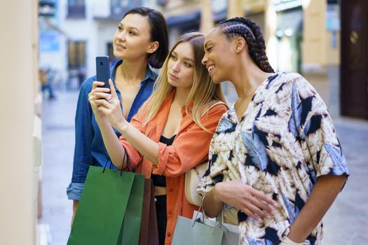 Side view of diverse female friends with shopping bags taking picture of showcase while standing near store on street during shopping