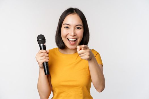 Enthusiastic asian girl with microphone, pointing finger at camera, suggesting you to sing, standing over white background.