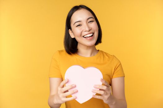 Romance and valentines day. Happy beautiful asian woman holding big heart card and smiling, standing over yellow background.