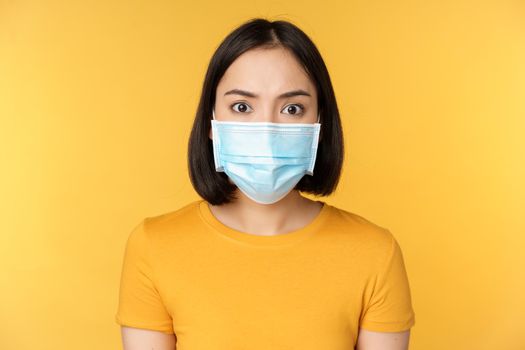 Portrait of shocked asian woman looking concerned and startled at camera, wearing covid-19 medical face mask, standing against yellow background.