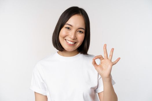 Everything okay. Smiling young asian woman assuring, showing ok sign with satisfied face, standing over white background. Copy space