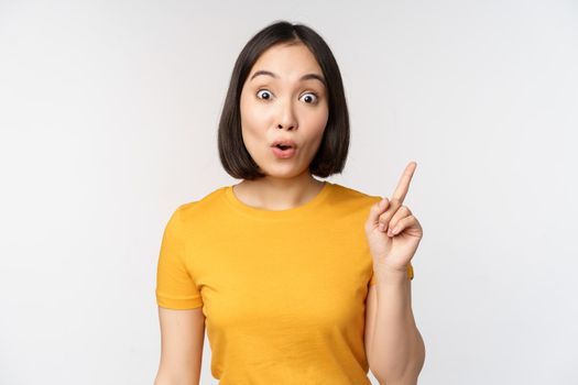 Beautiful young asian woman pointing finger up, smiling and looking amused at camera, showing advertisement, announcement on top, white background.