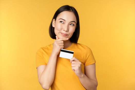 Thinking asian woman holding credit card and smiling, planning buy smth, standing thoughtful over yellow background.