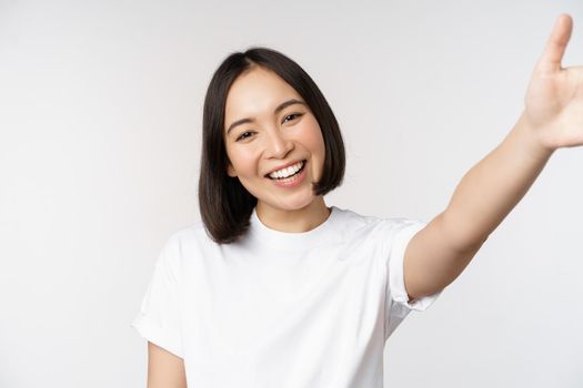 Beautiful young asian woman smiling, looking at camera, holding device, taking selfie, video chat, standing in tshirt over white background.