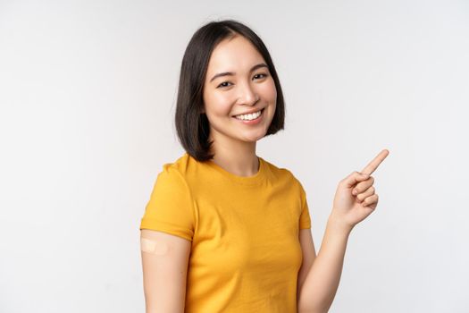 Covid-19 and vaccination concept. Smiling asian girl with band aid on shoudler, pointing finger at banner, showing vaccine campaign, standing over white background.