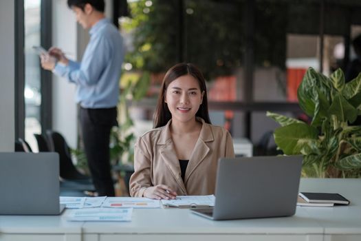 Portrait of young asian woman sitting at her desk with laptop while looking at camera in modern office