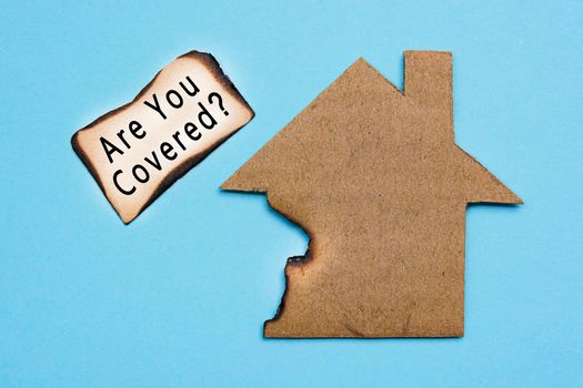 Are you covered text on burned paper house model on blue background. Home insurance concept. Flat lay. Directly above.