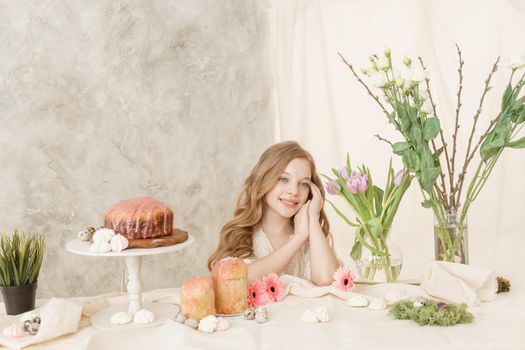 A girl with long hair in a light dress is sitting at the Easter table with cakes, spring flowers and quail eggs. Happy Easter celebration