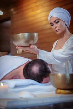 A woman performs Tibetan singing bowl therapy with a man lying under a white sheet. Relaxing meditation.