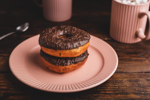 Stack of Homemade Chocolate Donuts and Mugs of Hot Chocolate with Marshmallow on Rustic Wooden Surface