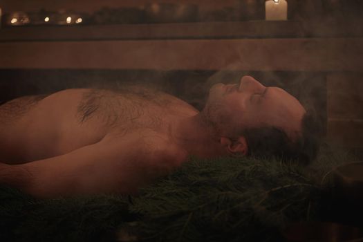 A man lies on spruce branches in a sauna, he is being performed a Tibetan ritual.
