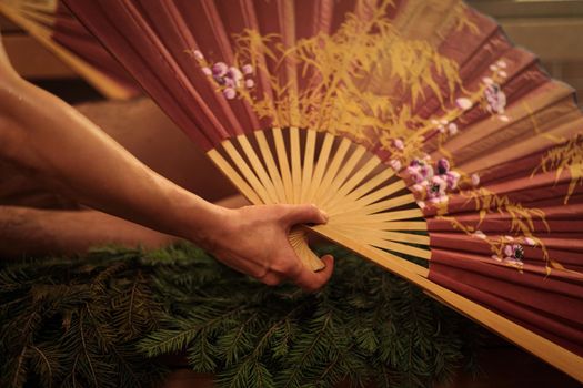 A woman performs a healing ritual with a Chinese fan for a man. A man lies on spruce branches.