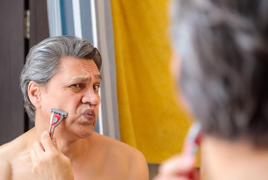An adult gray-haired man shaves with a razor in front of a mirror.