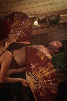 A woman performs a healing ritual with a Chinese fan for a man. A man lies on spruce branches.