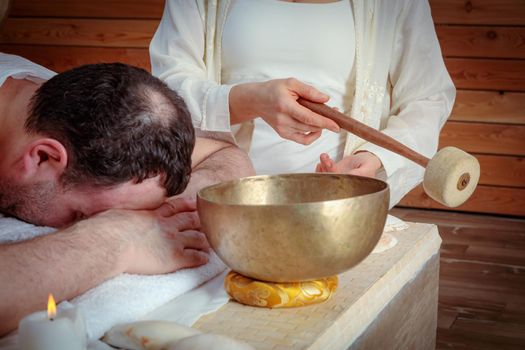 A woman performs Tibetan singing bowl therapy with a man lying under a white sheet. Relaxing meditation.