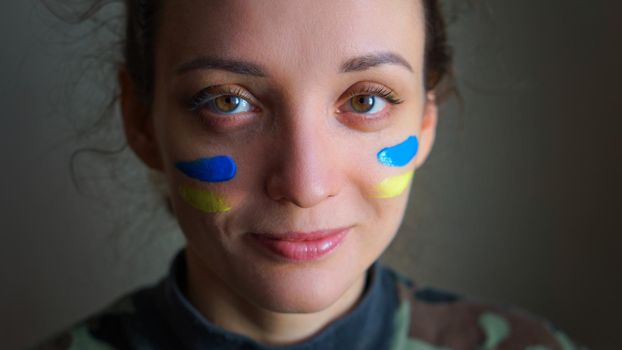 Indoor portrait of young girl with blue and yellow ukrainian flag on her cheek wearing military uniform, mandatory conscription in Ukraine, equality concepts.