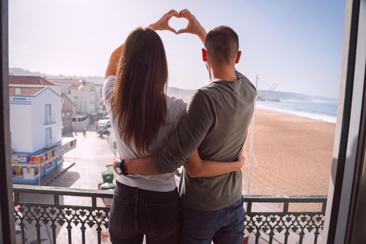 Young couple standing on the balcony and making heart shape with their hands. Couple in love hugging and spending valentine's day together. High quality photo