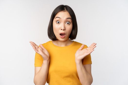 Close up portrait of asian woman looking surprised, wow face, staring impressed at camera, standing over white background in yellow t-shirt.