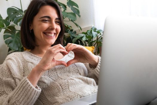 Happy young caucasian woman making heart shape with hands during online video call using laptop. Copy space. Love and technology concept.