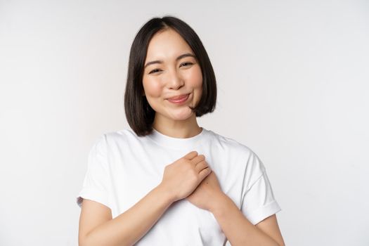 Portrait of carig young asian woman holding hands on heart, gazing with care and love, touched by something, feel flattered, standing over white background.