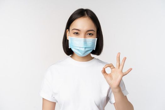 Covid-19, healthcare and medical concept. Happy asian girl smiling, wearing medical face mask, showing okay, ok sign, approve, recommending smth, white background.