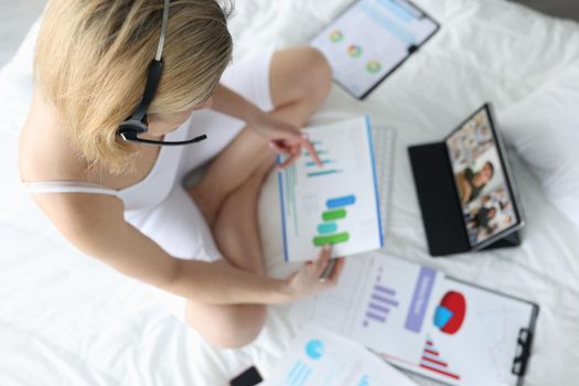 Woman with handsfree sitting on white bed with documents in hands and communicating via video link with colleagues top view. Home office concept