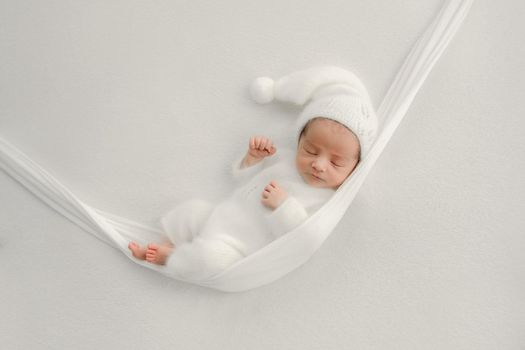 Newborn baby boy wearing white knitted costume sleeping in hammock. Infant child kid in festive holiday winter dress studio portrait