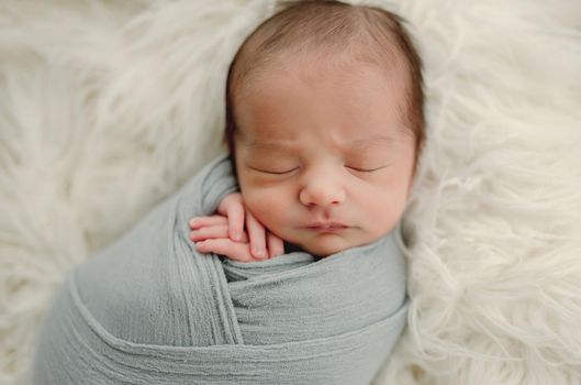 Newborn baby boy sleeping on white fur swaddled in fabric. Infant child kid napping studio portrait