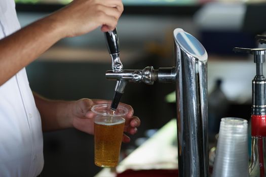 Close-up of bartender pouring beer from faucet machine in to big plastic glass. Order drink in pub, night club or bar. Alcohol, fun, party, service concept