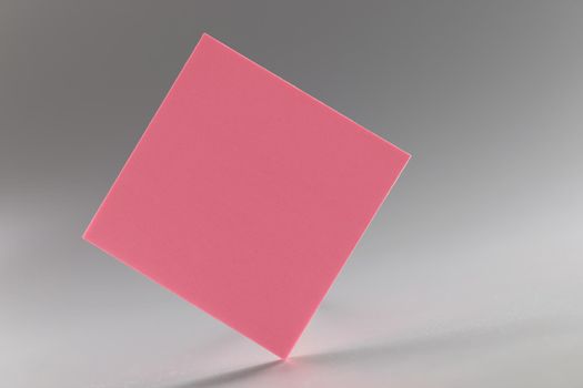 Close-up of pink square sticker standing on sharp edge on grey background. Piece of post it note in pink colour. Reminder, note, planning, sticker concept