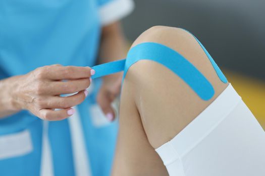 Close-up of physiotherapist apply kinesiology blue tape on client knee. Rehabilitation after injury, consultation in clinic. Physical therapy, cure concept