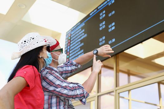 Portrait of couple going on trip during pandemic, flight delay, man point on wristwatch. Man and woman wear protective face masks. Virus, vacation concept