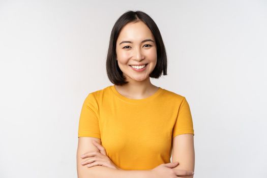 Portrait of young beautiful brunette, asian girl smiling and looking happy, cross arms on chest, standing over white background.