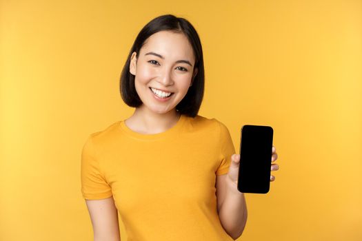 Beautiful happy asian girl showing mobile phone screen, application on smartphone gadget, standing over yellow background.