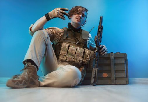 male soldier in camouflage is sitting by an ammunition crate on the floor with a weapon